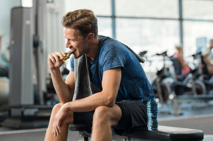 Smiling man in gym clothes sitting on a bench, enjoying a protein bar post-workout with towel over shoulder and water bottle nearby.